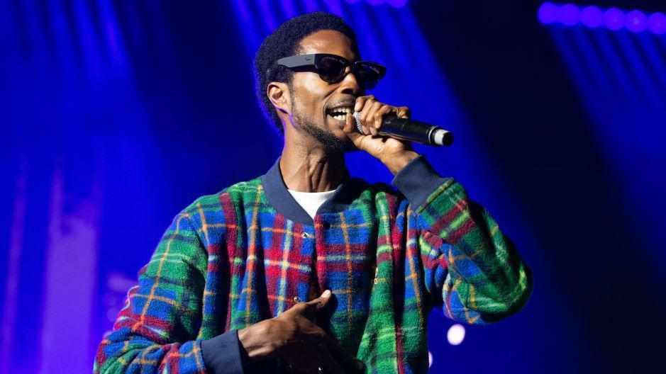 D Double E performs on day 1 of Reading Festival 2023. he is holding a black microphone, wearing dark sunglasses, and has a green, blue and red striped sweater top on. The background is of dark blue coloured lights.