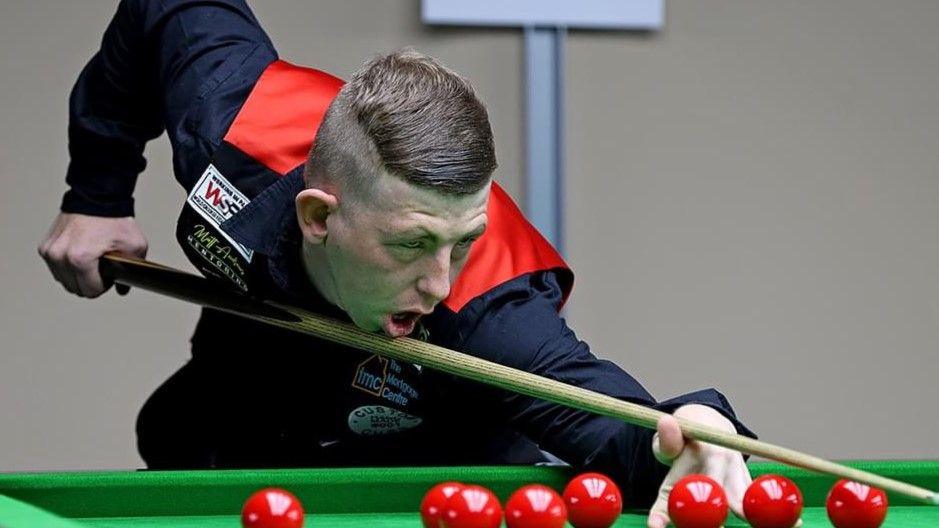 David Church playing snooker while wearing a red waistcoat and black shirt. Under a bright light, he is leaning over the green felt of a table, his cue at the ready, concentrating on a shot.