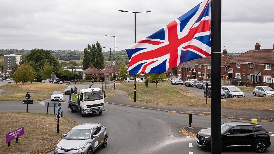 A Union Jack flag hangs from a lamppost above a roundabout, as cars pass beneath it and houses and other flags hanging from lampposts can be seen in the distance.
