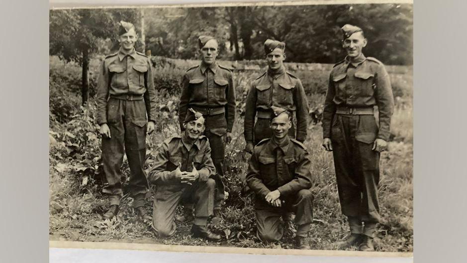 Six soldiers in uniform during World War Two. Two men are kneeling down while four others stand in a row behind them as they pose for the photograph. Vera Parnaby's father is kneeling on one knee in the bottom left. He is smiling and has his fingers interlocked with his thumbs sticking up.