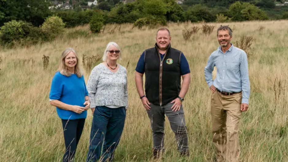 Members of the society and Moreton Cullimore, smiling, whilst standing in an overgrown field with woodland in the background