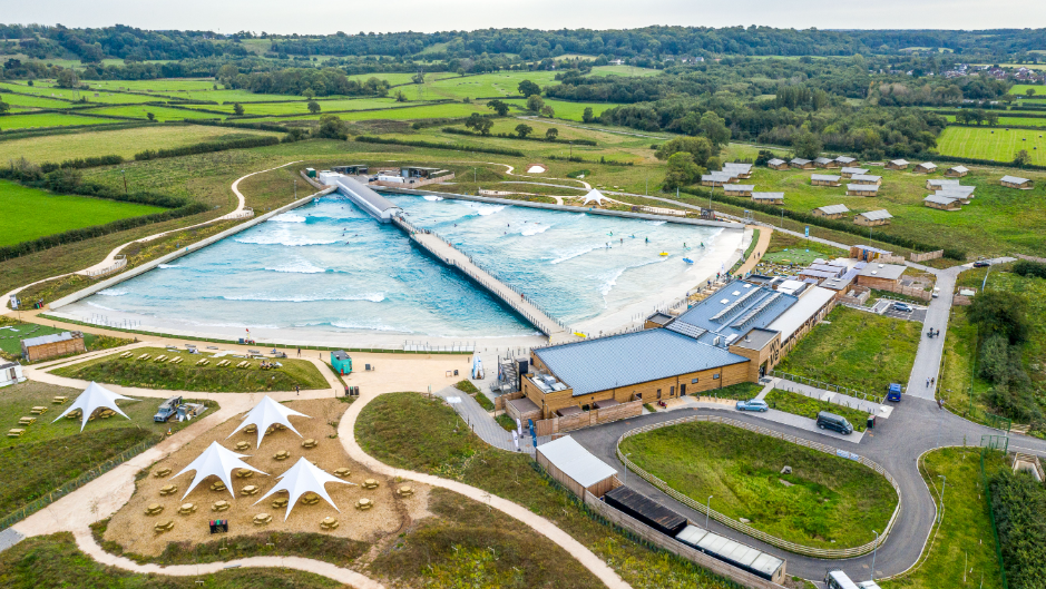 An aerial image of The Wave in Bristol. It is a large triangle shape with a pier extending through the middle of it. It is surrounded by fields and trees, with a small roundabout in front and a wooden reception area and restaurant.