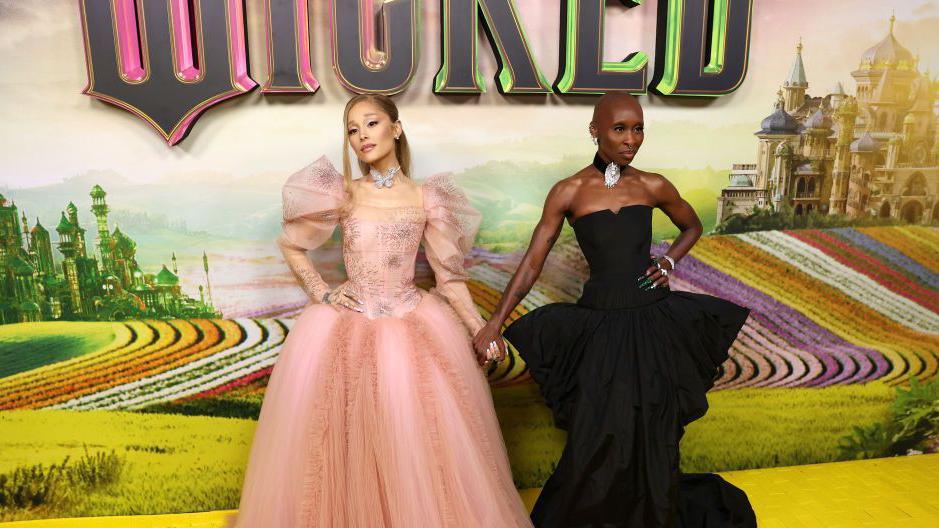 The image shows two stars of the new Wicked movie - Ariana Grande and Cynthia Erivo. Ariana Grande wears a pink dress and Cynthia Erivo wears a black dress. 