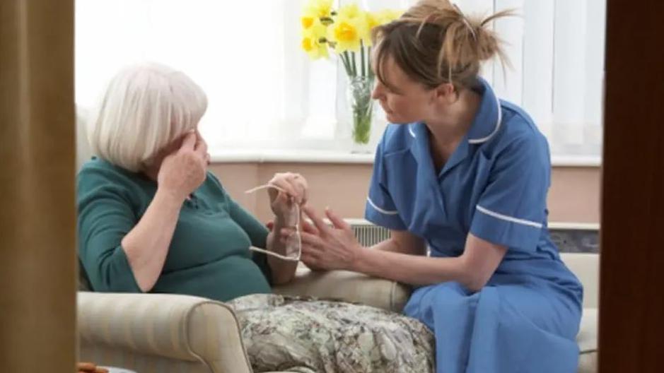 Generic photo of a social care worker wearing a typical blue uniform and an elderly woman wearing a green jumper and a skirt. She is holding her glasses and appears to be crying 