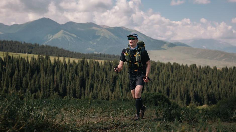 A man in a t-shirt, shorts, hat and sunglasses hiking on a mountain in Kyrgyzstan.