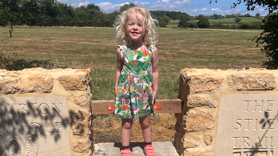 A little girl with blonde hair standing on a stone stile with the words 'THE STILE TRAIL' carved into one side. There is an open field of grass behind her, with trees in the distance