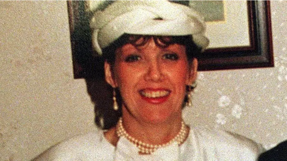 A woman smiling at the camera. She is wearing a white dress and hat. She is wearing pearl earrings and necklace. She is standing in front of a framed picture hanging on a wall.