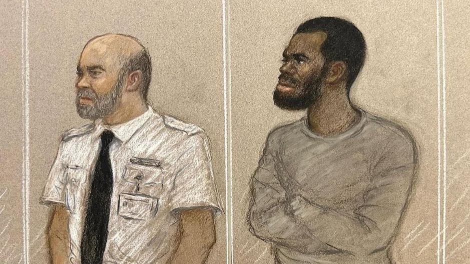 A court sketch of a man in a grey tracksuit standing in the dock with his arms crossed. He has short, dark hair and a beard and moustache. Next to him is a dock officer in a white shift and black tie.
