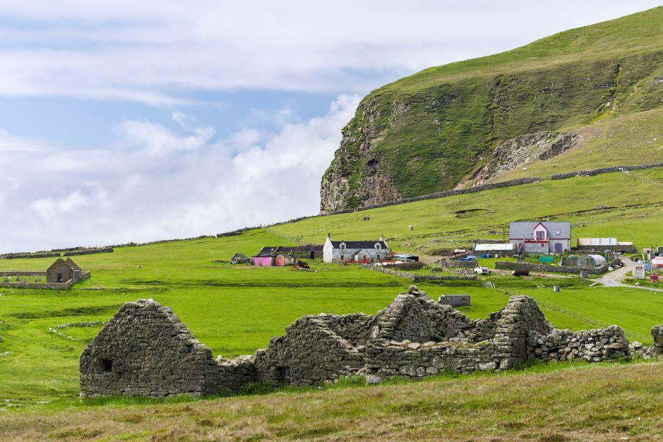 Remote farmhouse on the Shetland island of Foula, with ruins of a farm building in the foreground, amid grassy fields, and rocky high cliffs in the background.