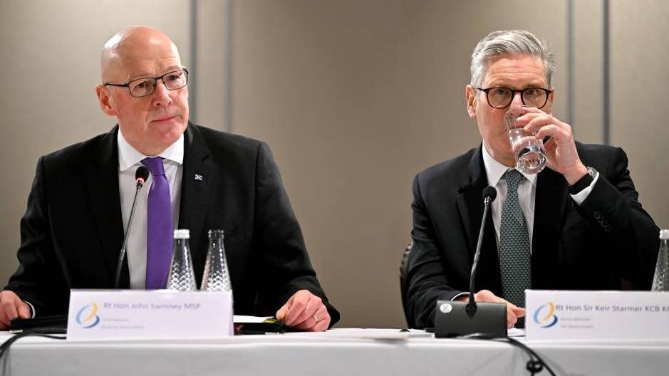 John Swinney, wearing a dark suit and a purple tie sits at a table with a placard showing his name. There is also a microphone and two bottles of water in front of him. To his right is Keir Starmer, who also has a microphone and placard in front of him. He is wearing a dark suit and green tie. He is taking a drink from a glass of water.