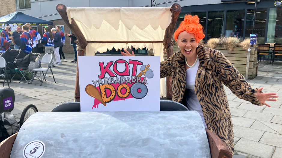 A woman is standing next to one of the karts. She is wearing a beige and black animal print coat, a white dress, and an orange wig. The kart has a silver barrel at the front and a sign. 
