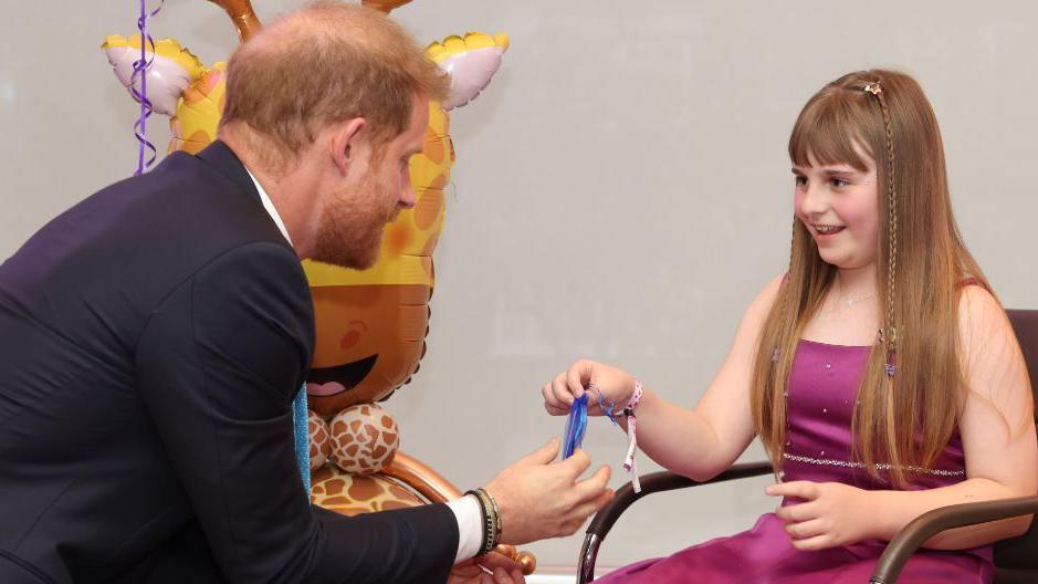 Prince Harry sits on the left wearing a dark blue suit, with a white shirt and sky blue coloured tie. He is talking to Scarlett, who is 10 years old, she sits in a dark brown leather chair wearing a purple silk gown, which has diamond embellishing on the top. Scarlett had light brown long hair with two plaits, fastened by star clips that framing her face and a fringe. Scarlett is smiling while talking to Prince Harry and there is a helium inflated giraffe balloon in the background.