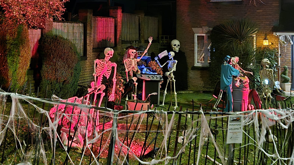 A house lit by red lights with skeletons decorating the garden in various poses including around a barbecue. A fence in the foreground is decorated with fake cobwebs