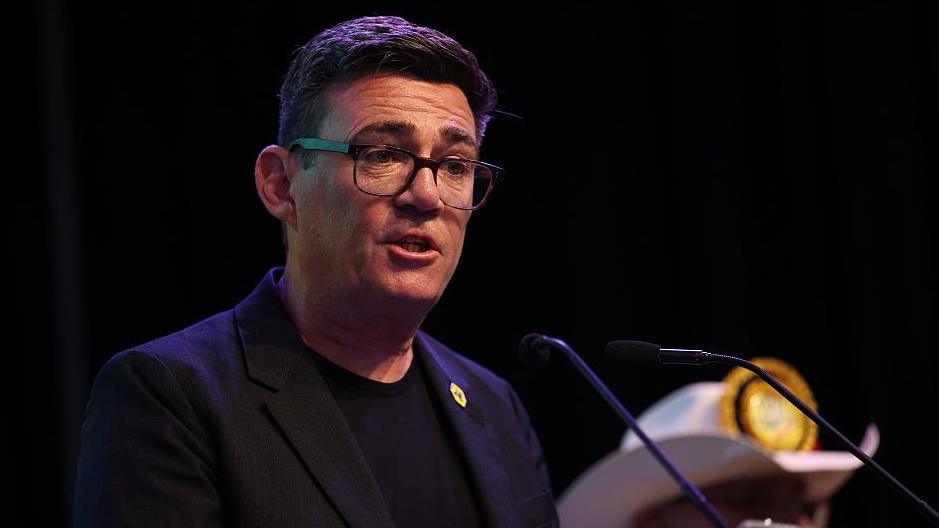 Andy Burnham speaking in to microphone while standing at podium. He is wearing a black suit jacket on top of a black t-shirt