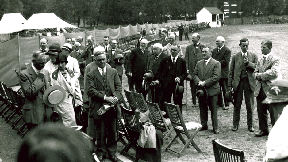 A large group of smartly dressed people, mostly men, walk towards wooden seats at the County Ground. The seats have all been placed around the circular pitch. The picture is black and white.