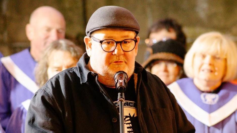Comedian Johnny Vegas wearing a black cap, black glasses, black shirt and black and white t-shirt stands in front of a microphone with people blurred out in the background dressed in purple.