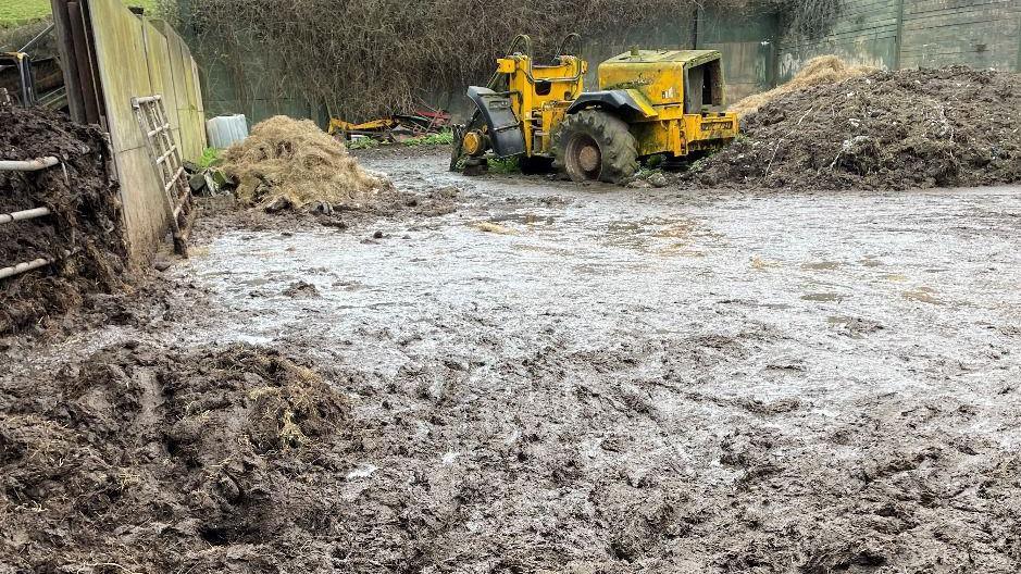 The image shows a large amount of animal slurry in a farmyard with piles of straw or hay and wat appears to be more slurry along with the remains of a wrecked heavy machinery coloured yellow. There is a wooden fence to the left and a metal fence which is apparently holding back a pile of animal waste.