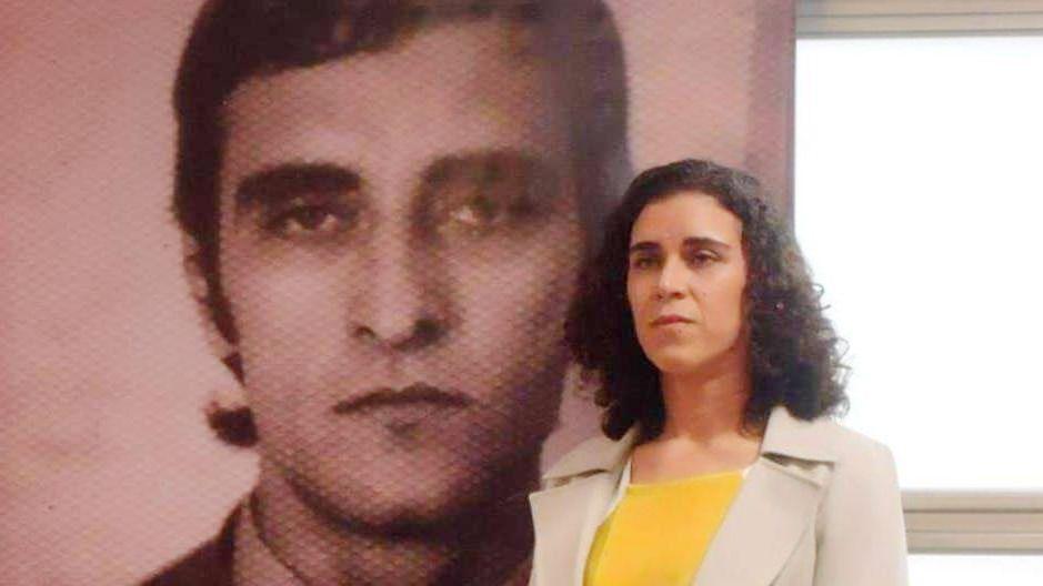 Tessa, a Brazilian woman wearing a yellow top, stands in front of a picture of her father