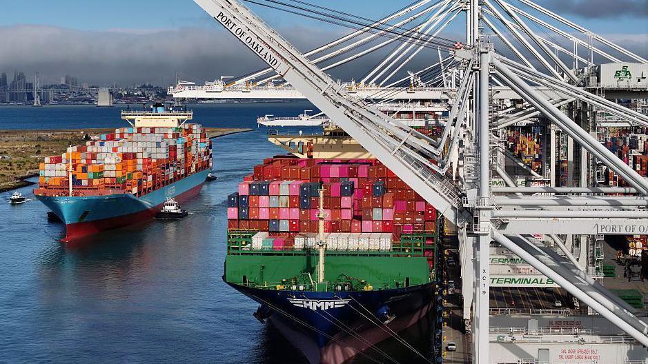 Two ships with multi-coloured containers at the Port of Oakland, California. One of the ships is tied to the dock while the other is being escorted by two pilot boat.