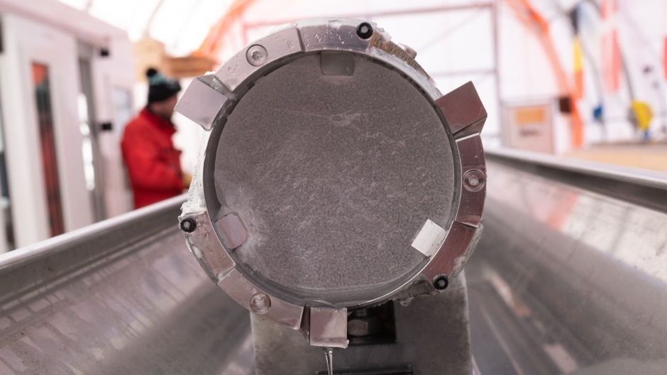 The end of an ice core in a metal tube. It is inside a large tent in Antarctica where the ice cores are being kept. On the left-hand side, there is a person wearing a bright red jacket and a bobble hat