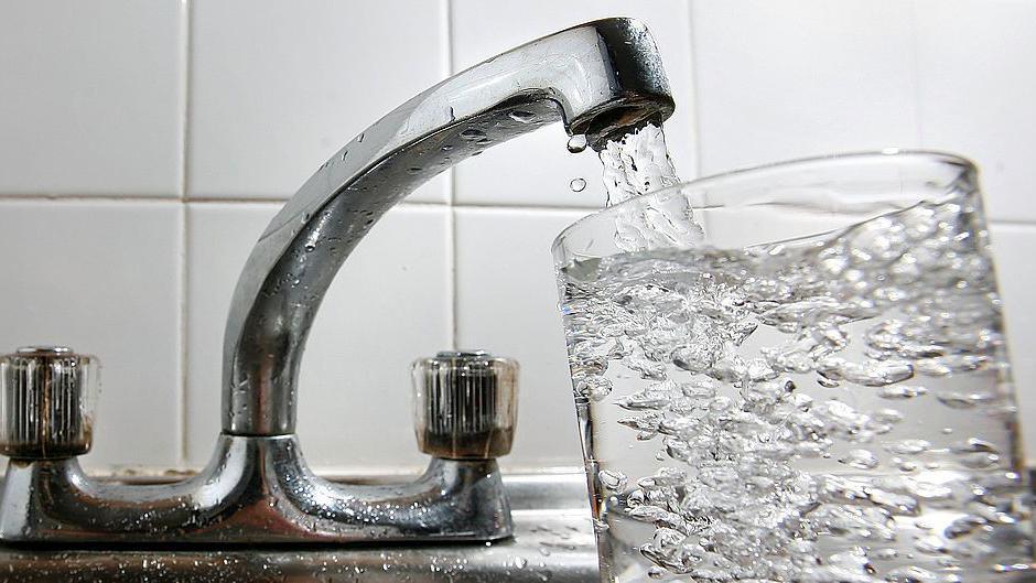 A glass of water is being filled from a silver tap. Water can be seen coming out of the spout and filling up a clear glass.