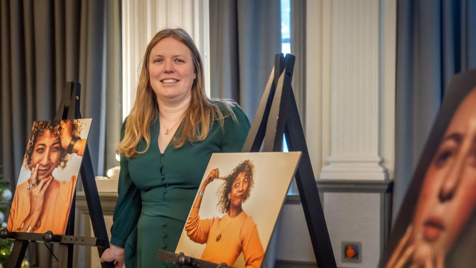 A woman with long blonde hair wearing a green dress smiles at the camera. She is standing next to easels showing photographs of a woman in orange.