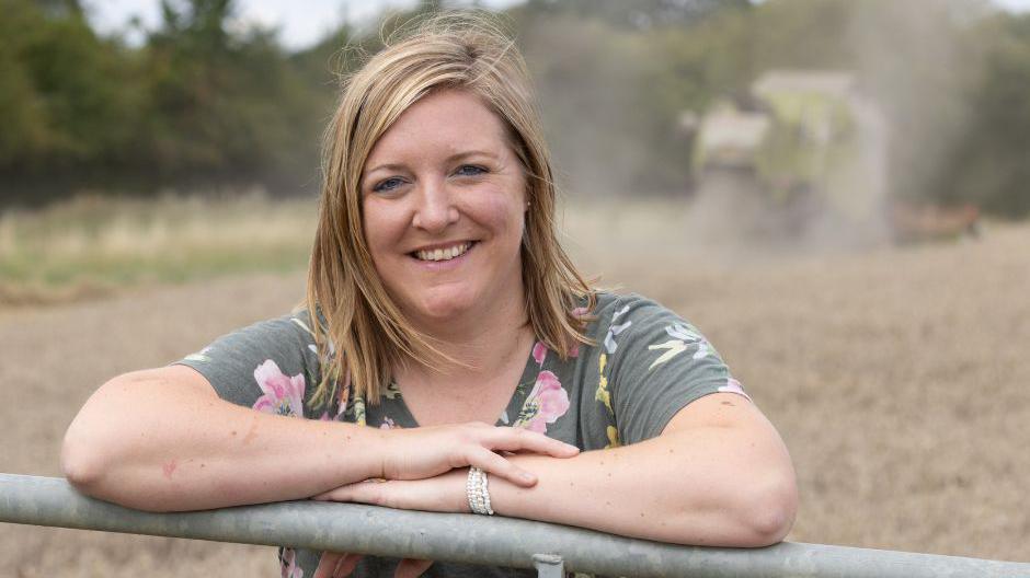 Milly Fyfe in a grey dress with flowers on it stands in a field leaning with her arms on a metal gate and smiles at the camera.