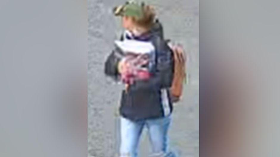 A grainy CCTV image of a woman looking away from the camera, wearing a green cap, brown rucksack and brown jeans. She's carrying an unidentifiable object in her arms.