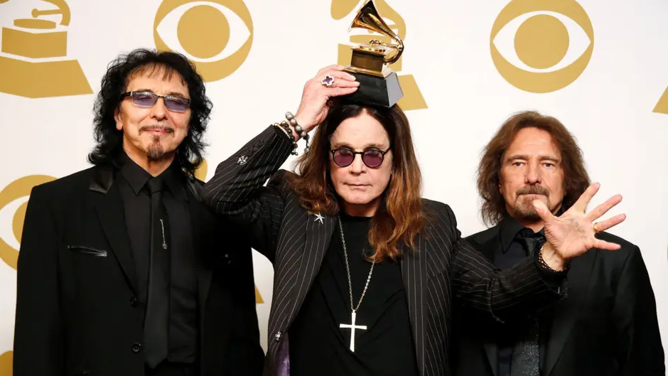 Ozzy stands in the centre between two bandmates and has an award resting on his head. The trio are all dressed in black. Ozzy has a cross necklace on and is wearing shades.