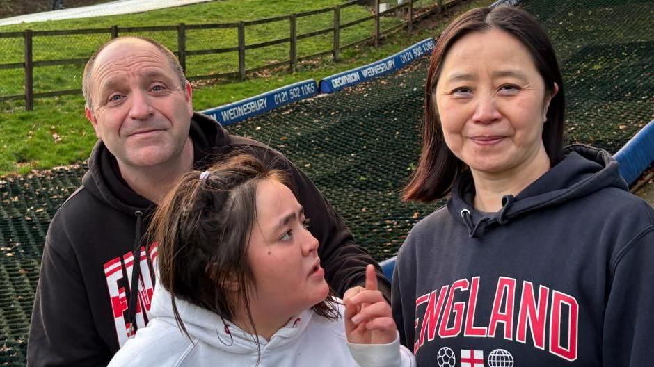 There are three people called Simon, Mandy and their daughter Emma. The woman on the right (Mandy) is wearing a dark grey hoodie with the word England written on it with big red letters. She has dark brown shoulder-length hair. In the middle, there is a young woman (Emma) looking up towards her mum. She is wearing a white jumper and has long dark brown hair. The man on the left (Simon) is wearing a black jumper with red and white lettering. The letters EN are visual. He has a grey hair in a buzz-cut hairstyle and is balding.