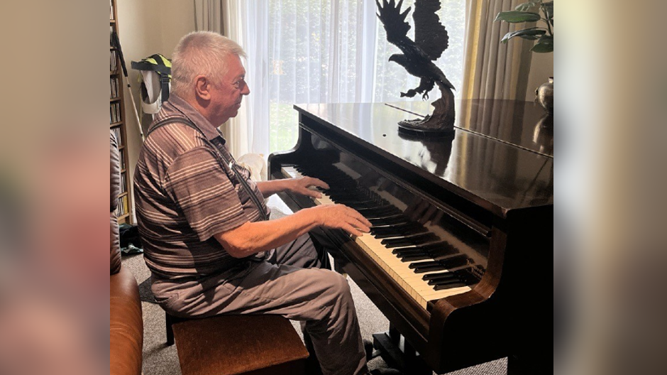 Graham Grange is sat at the piano, with his hands resting on the keys as if he is playing it or about to. There is what appears to be a wooden model of an eagle on top of the piano