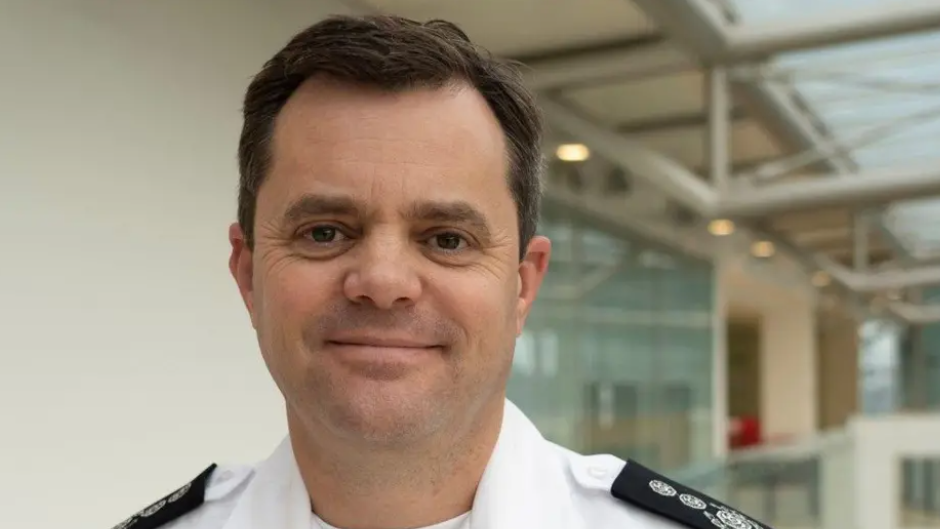 A man with greying brown hair and brown eyes looks directly into the camera with a half smile on his face. His white collared shirt has black fire service epaulettes.