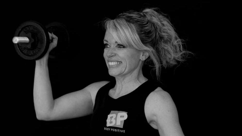 Loretta Davies pictured lifting a dumbell and smiling away from the camera. The photo is in black and white and she wears a "body positive" vest. 