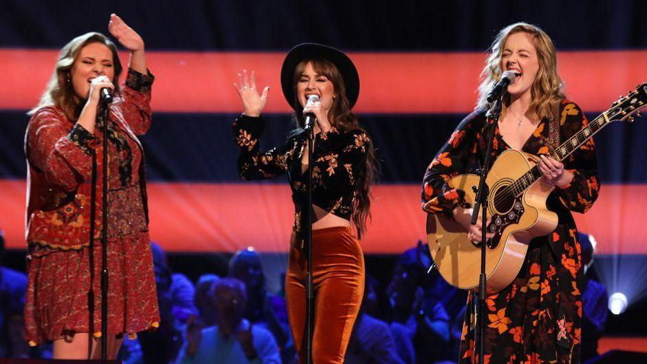 Three women, one holding a guitar, singing into microphones in front of a studio audience bathed in blue light.