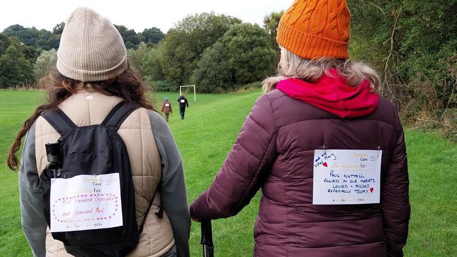 Two women stand side by side in a field, with their backs to the camera. On their backs they have large postcards where they have written the names of loved ones whom they have lost.