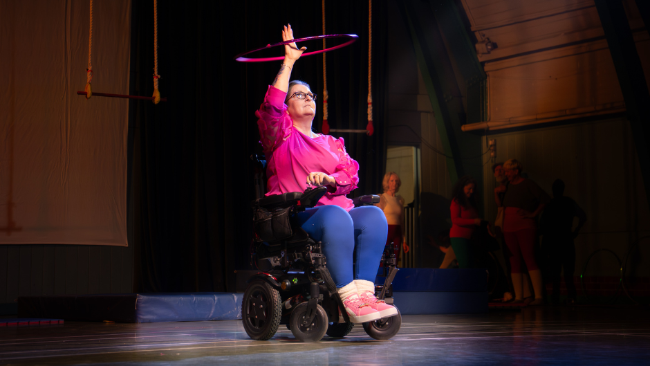 Claire sits in her wheelchair on stage, while spinning a hula hoop over her head. She wears blue leggings and a bright pink top.