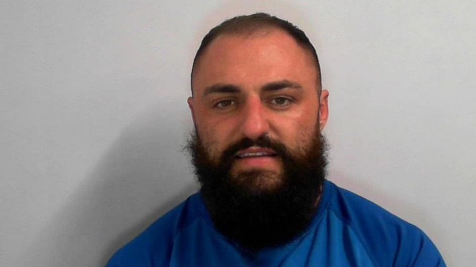 Macauley Nesfield with a thick black beard and black hair and wearing a blue top, stood in front of a grey background