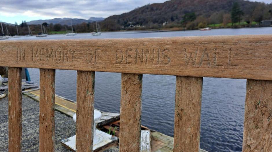 Close up of a wooden bench on the pier of a lake. It has the words 'IN MEMORY OF DENNIS WALL' inscribed along the top. 