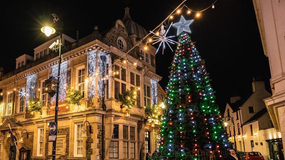 The municipal building illuminated in Christmas lights with a lit up green Christmas tree to the right.