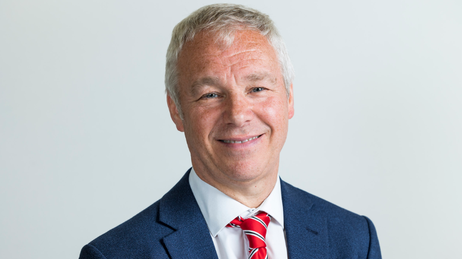 Councillor John Ennis smiling at the camera, wearing a white shirt, blue blazer and a red tie.