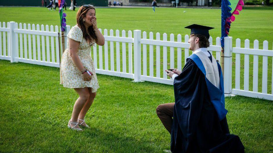 Two people, one in a dress standing up with her hand over her mouth, the other in a graduation gown on one knee proposing on the grass in front of a white picket fence