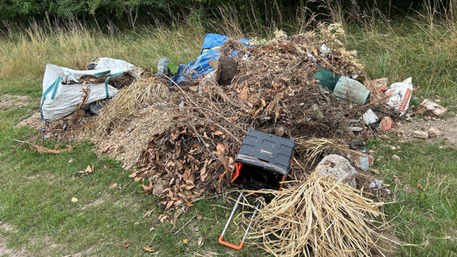 A large pile of fly-tipped waste including a small plastic shopping trolley, dried grass, leaves and a garden pot.