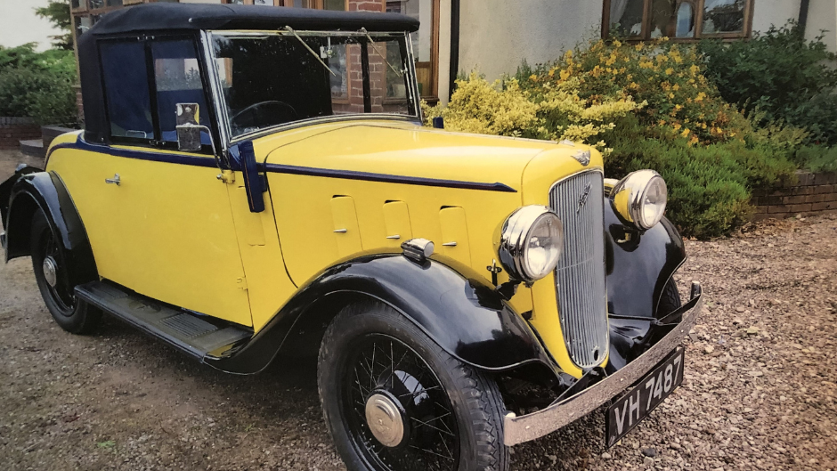 A restored yellow classic car with a black roof parked on the gravel outside a care home. The building is behind the car and bushes are visible to the right.