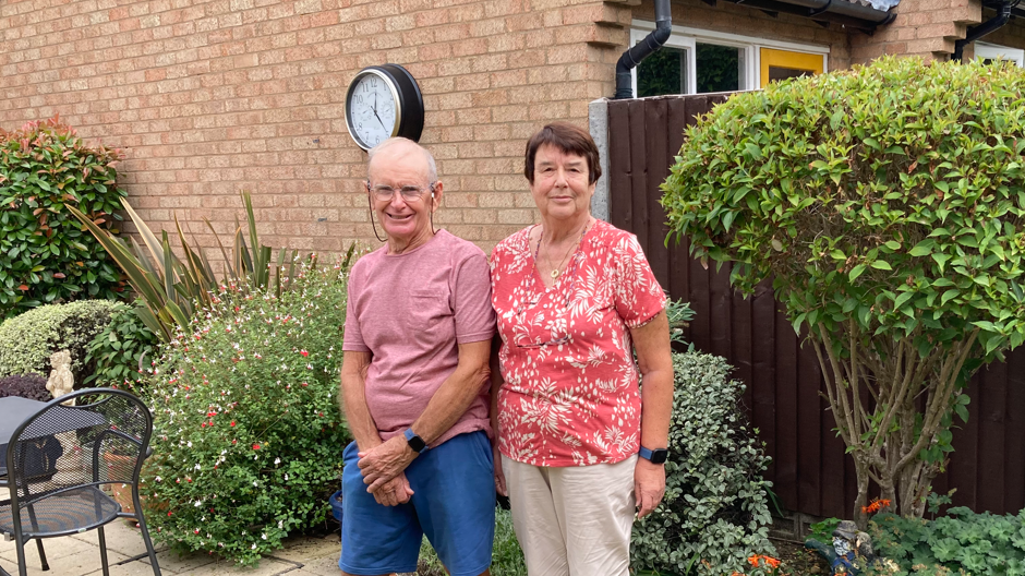 Pete and Sue Woolfitt are standing next to each other in a garden with bushes and greenery blooming behind them. They are both smiling and wearing red tops. Pete is also wearing glasses, blue trousers and is clasping his hands in front of him. Sue has short brown hair and is wearing cream-coloured trousers.