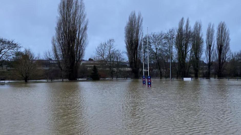 Flooded rugby pitch. A rugby goal is at one end with tall trees at the edge of the field.