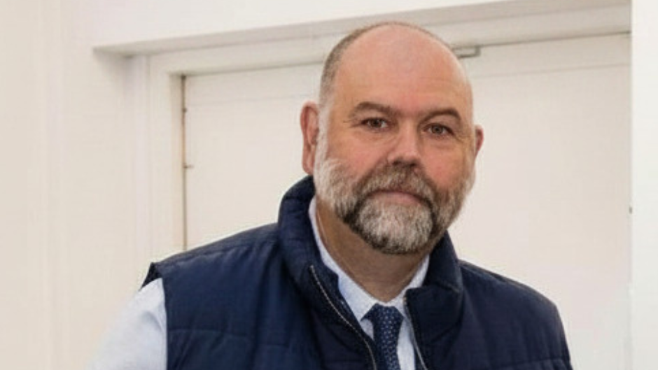 A man with a beard looking at the camera. He is wearing a navy sleeveless jacket, a white shirt and a dark blue tie.