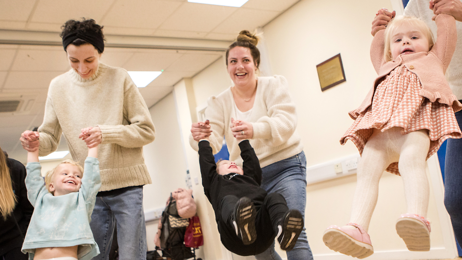 Three women, laughing and smiling as they swing their young toddlers by their arms in a village hall