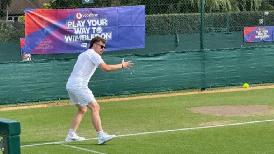 Matthew Brough wearing white shorts and a white t-shirt and white trainers, bent over with one arm posed in a position that suggests he has just hit a tennis racket. He is stood on a tennis court.