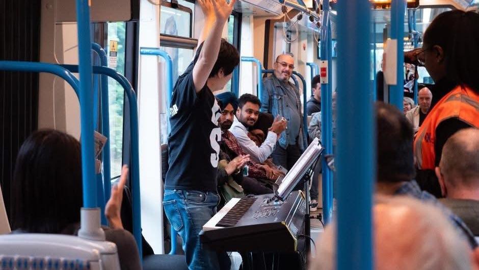 A person raises their arms, as they stand in front of a large keyboard, which appears to be on a tram or train. There are people looking on