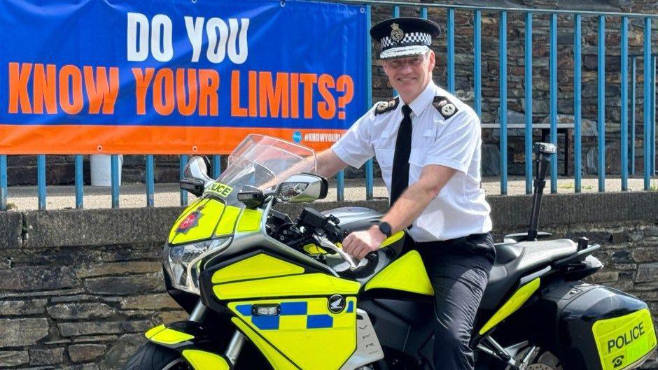 Russ Foster, in police uniform, sitting on a Isle of Man Constabulary yellow bike in front of blue railings and a poster saying 'do you know your limits?'